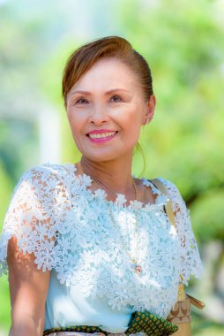 Orta yaşlı, güzel, sağlıklı bir Asyalı kadının ön portresi Tayland tarzı giyinmiş yeşil ağaç arka planında mutlu bir şekilde gülümsüyor.