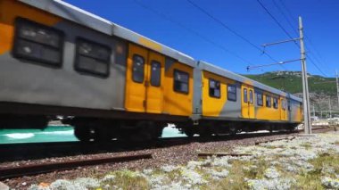 Cape Town, Güney Afrika - 08 Kasım 2019: Arka planda uçurtma sörfçüsüyle kıyı şeridi boyunca hareket eden bir yolcu treninin hızlandırılmış videosu 
