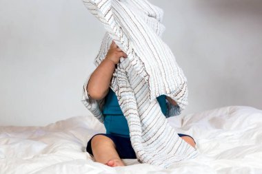 Beyaz yorganın üzerinde çizgili bir battaniyenin altında oynayan beyaz bir çocuk.