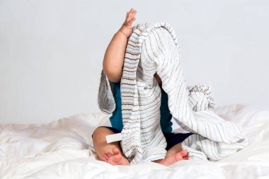 Beyaz yorganın üzerinde çizgili bir battaniyenin altında oynayan beyaz bir çocuk.