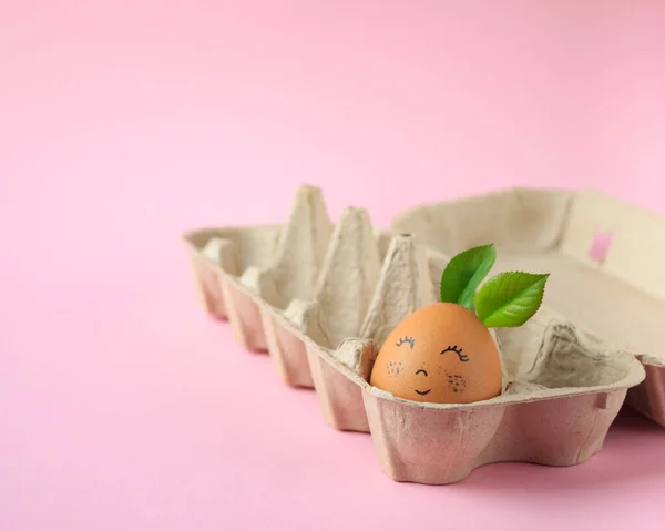 a chicken egg with a cute painted face on it, lying in an egg box on a pink background, a festive Easter concept