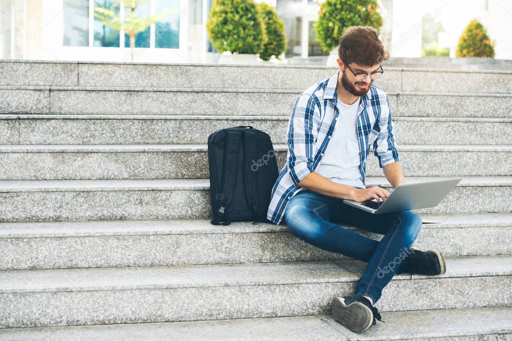 Student man sitting on stairs outdoors and working on laptop