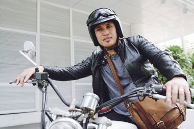 Asya motorcu açık havada vintage motosiklet üzerinde otururken kask ve deri ceket giyiyor düşük açılı görünümü