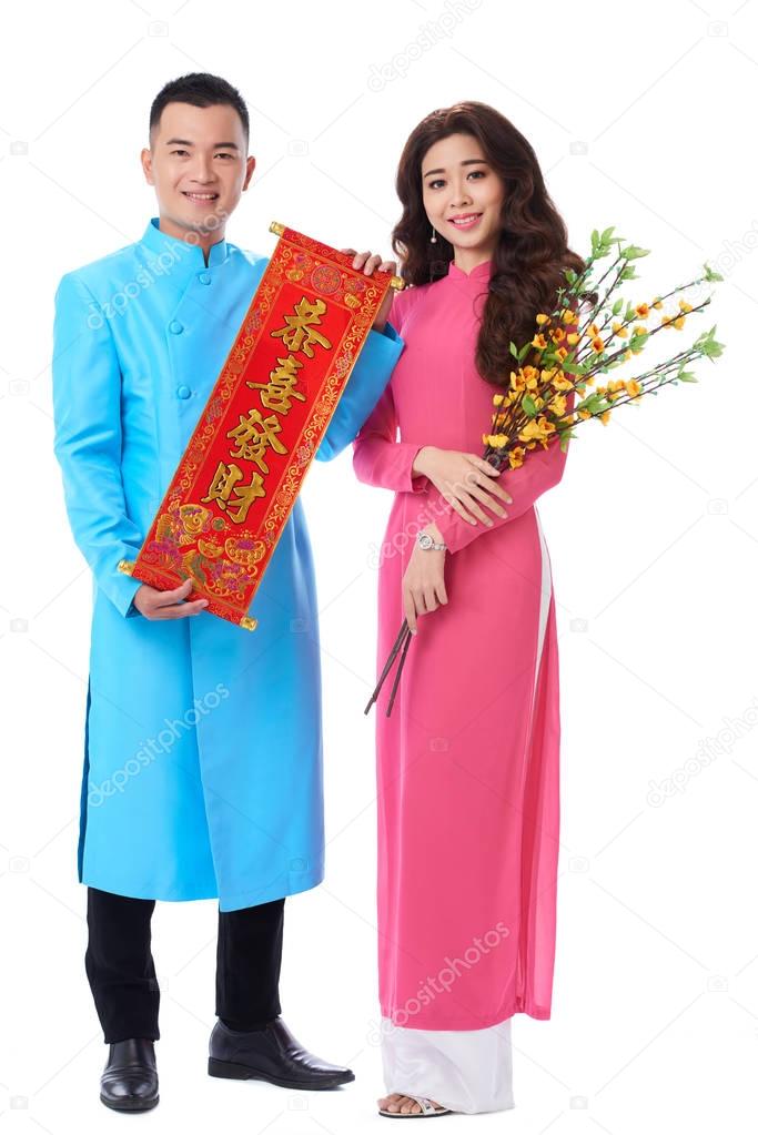cheerful Vietnamese couple wearing traditional costumes and holding Lunar New Year attributes in hands