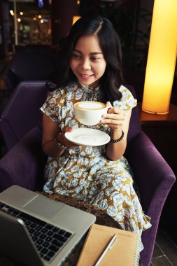 Genç kadın zevk büyük bardak kahve ve bir dizüstü bilgisayar ekranında okuma gülümseyerek