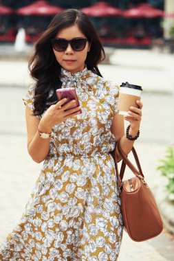 Şık iş kadını eve servis kahve ile açık havada yürüyüş ve onun smartphone haber okuma güneş gözlüğü