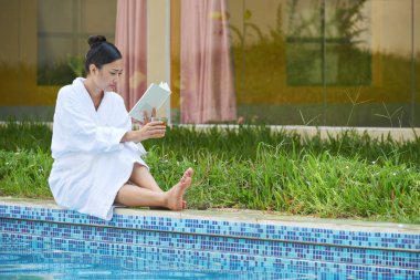 Havuz kenarında otururken, bir kitap okuma ve bardak suyu içme genç ve güzel kadın