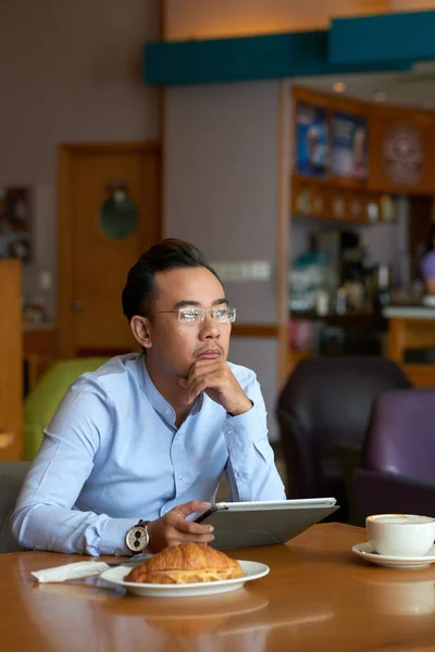 Pensive Asian entrepreneur with digital table sitting at cafe table