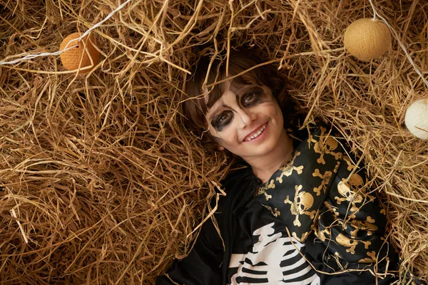 Smiling boy with scary Halloween make-up lying in hay
