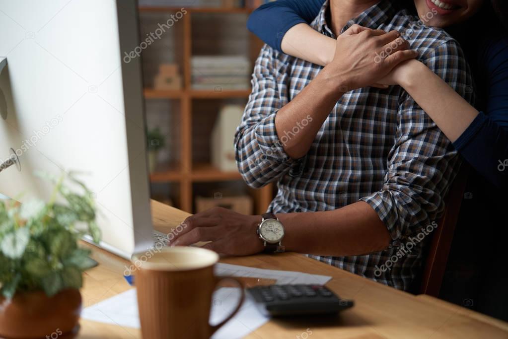 Woman hugging her husband from behind when he is sitting at workplace