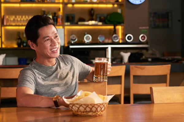 Happy middle-aged Vietnamese man drinking beer and watching match in sports bar
