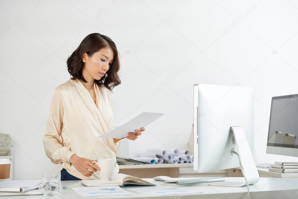 Portrait of young Vietnamese businesswoman reading document standing near office desk