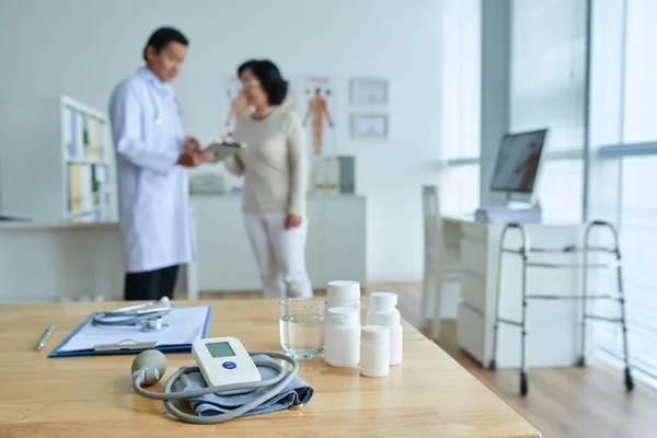 医生办公室的木质办公桌 药瓶和血压监测仪 亚洲医生和他的高级病人站在距离和谈话 — 图库照片