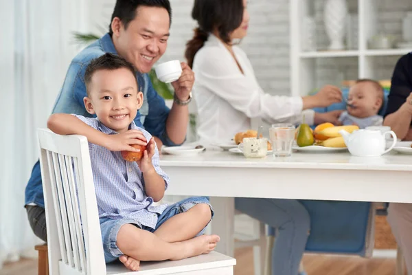 幸福的亚洲男孩的画像微笑对照相机 藏品苹果 当坐在早餐桌与家庭 — 图库照片