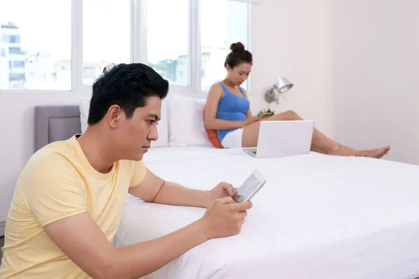 忧郁的亚洲年轻人发短信 他的妻子坐在智能手机和笔记本电脑的背景 — 图库照片
