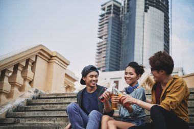 Happy Asian friends sitting on shabby stairs and celebrating their long-awaited gathering with soft drinks, portrait shot clipart