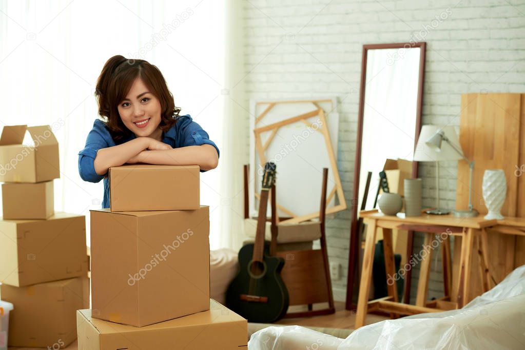 beautiful young Asian woman in room leaning on carton boxes and looking at camera