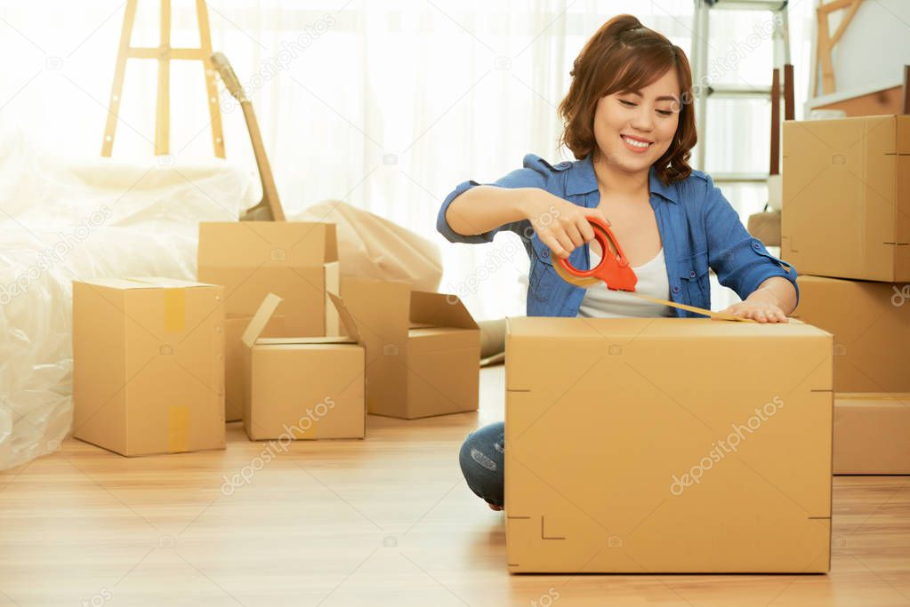 asian young woman packing stuff in carton box with Scotch tape for moving and sitting on floor 