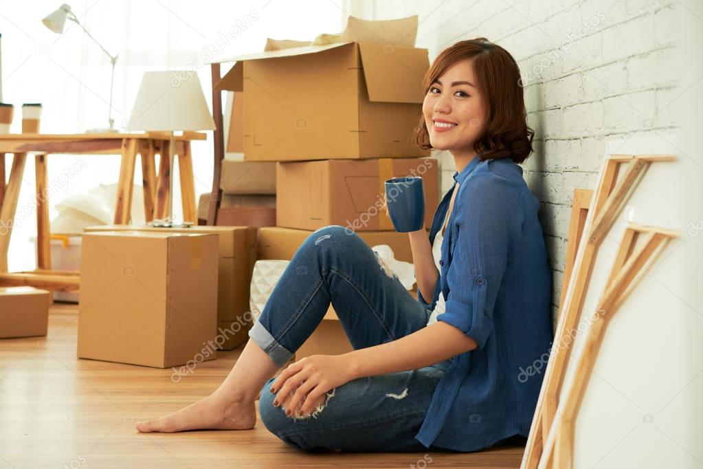 beautiful young asian woman sitting on floor in room and holding tea cup, looking at camera 