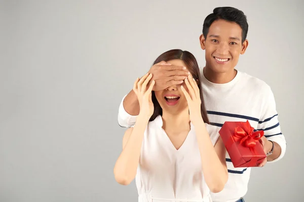 英俊的亚裔男子穿着条纹套衫给他的女朋友带来惊喜 他拿着礼物盒 用手遮住她的眼睛 灰色背景 — 图库照片