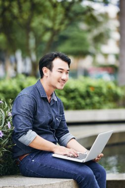 Neşeli Vietnamca genç adam açık havada oturma ve laptop e-postaları okuma