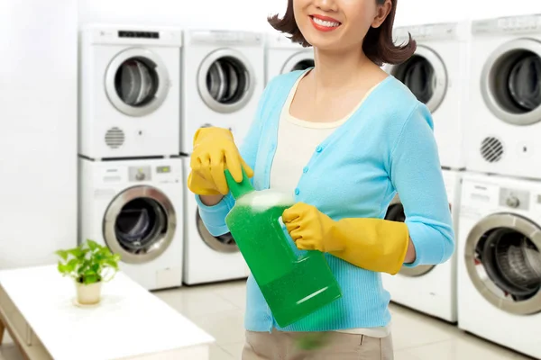 微笑的少妇佩带的橡胶手套打开瓶液体洗涤剂 当站立在宽敞自助洗衣店时 — 图库照片