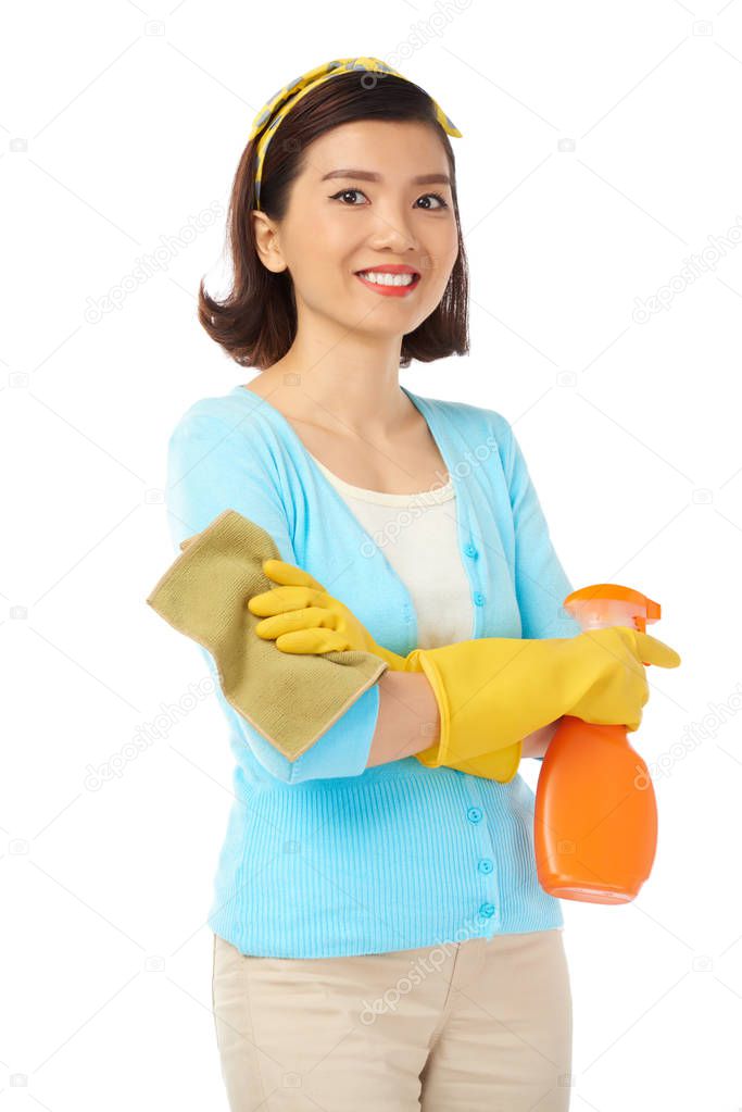 Asian housewife looking at camera with toothy smile while holding rag and spray cleaner in hands, isolated on white background