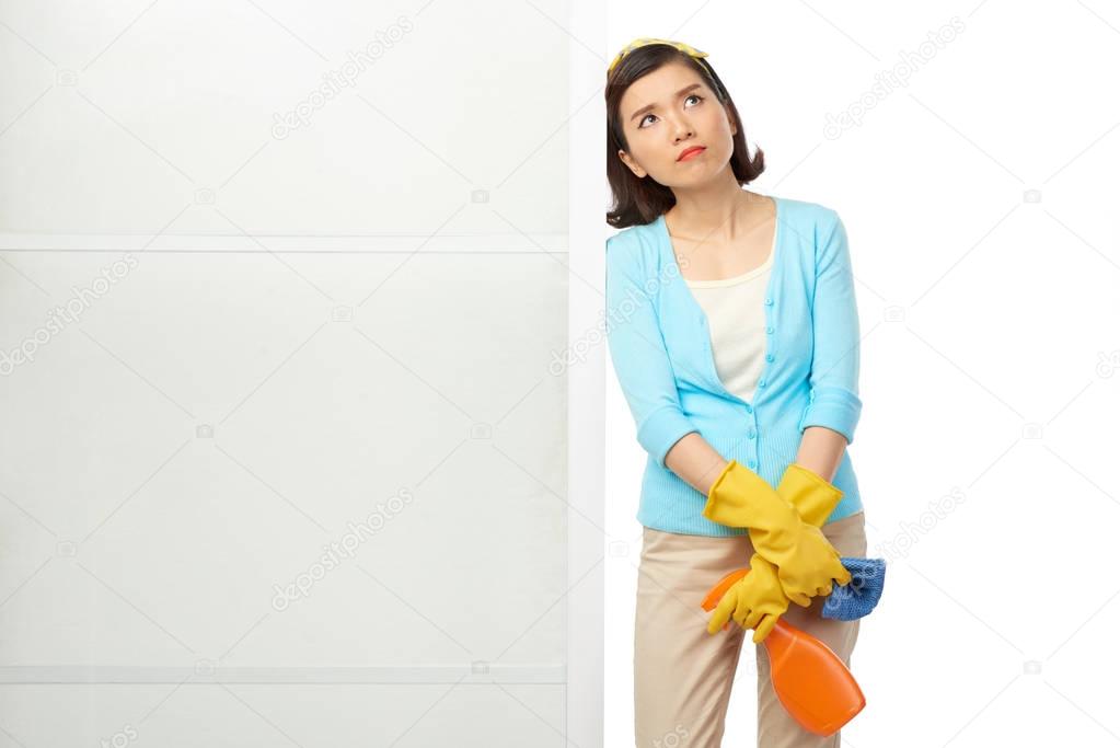 Pretty Asian housewife with detergent and rag in hands looking away thoughtfully while leaning on white wardrobe