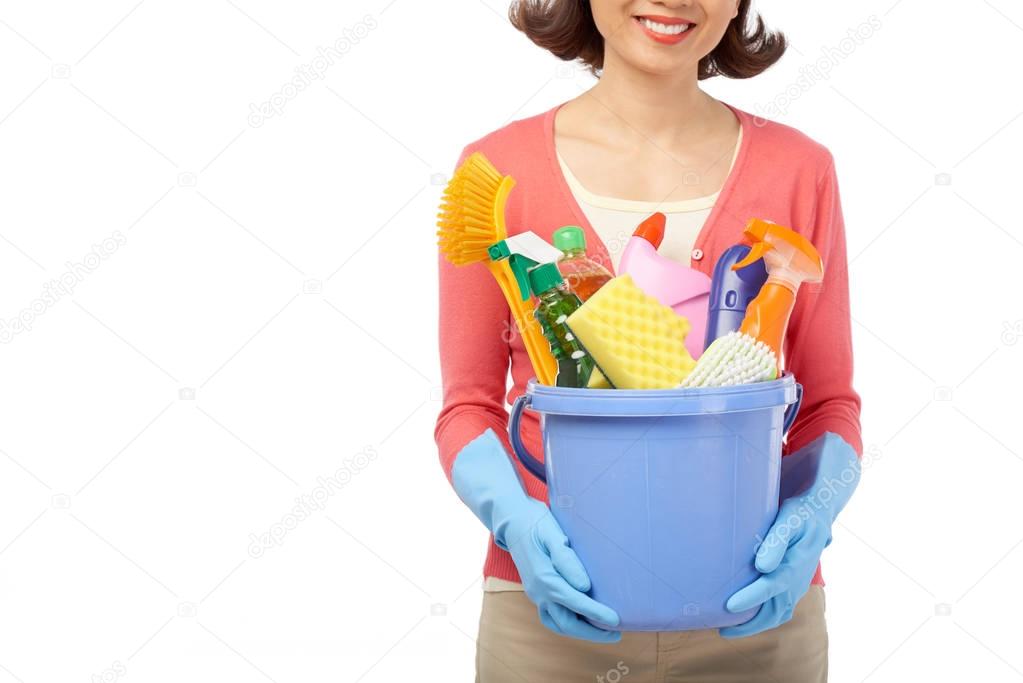 smiling housewife holding bucket with cleaning utensils in hands while standing against white background