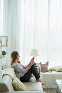 Kanepede oturan ve bir tablet bilgisayarda okuma genç kadın yan görünüm