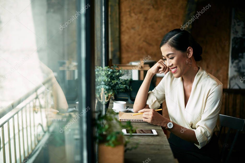 Cheerful business woman taking notes in notebook when having coffee in cafe