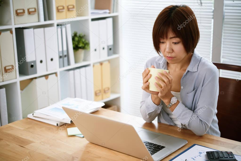 Asian business woman with cup of coffee in hands reading something on laptop screen