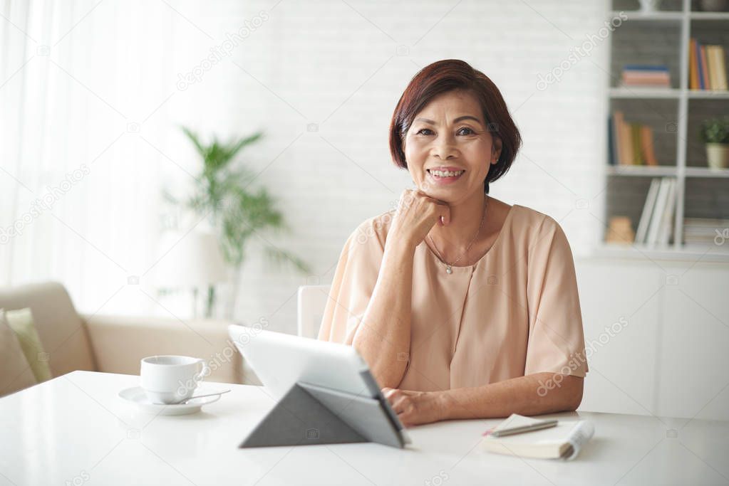 Portrait of cheerful senior business lady sitting at table with digital tablet and coffee cup