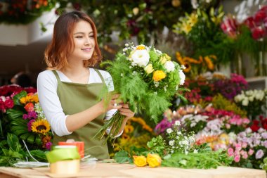 Asian florist wearing apron standing at counter and making bouquet for client, interior of flower shop on background clipart