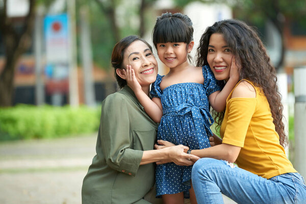 Family portrait of attractive middle-aged woman and her mature mother sitting on haunches while embracing cute little girl 