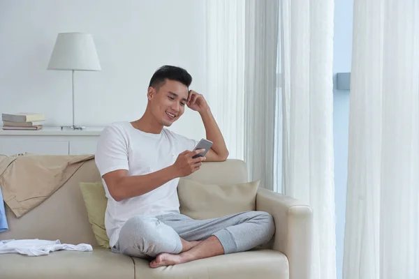 Guapo Vietnamita Joven Sentado Sofá Leyendo Algo Divertido Teléfono Inteligente — Foto de Stock