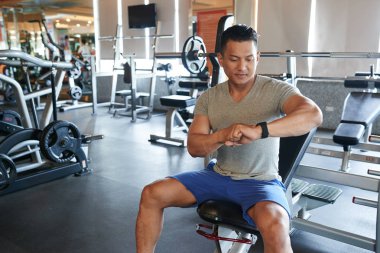 Asya genç sporcu spor salonunda bankta oturmuş ve fitness İzleyicisi'ni ayarlama