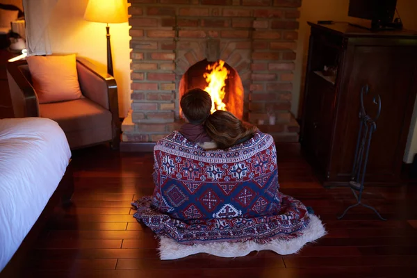 Young couple under warming blanket sitting at fireplace on cold winter evening