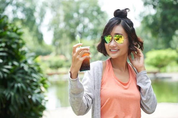 亚洲女子戴墨镜 手持塑料杯冰茶 背景绿色公共公园 — 图库照片