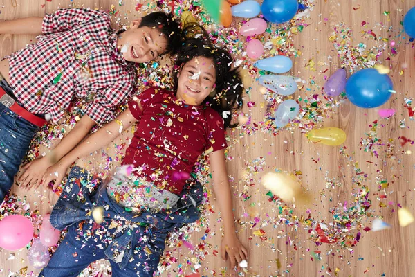 Happy twins lying on the floor with sparkling confetti and balloons around