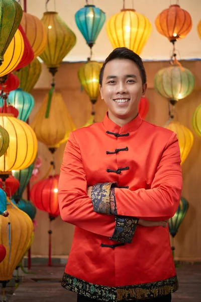 Confident Asian man wearing traditional costume standing with arms crossed while posing for photography, Chinese lanterns on background