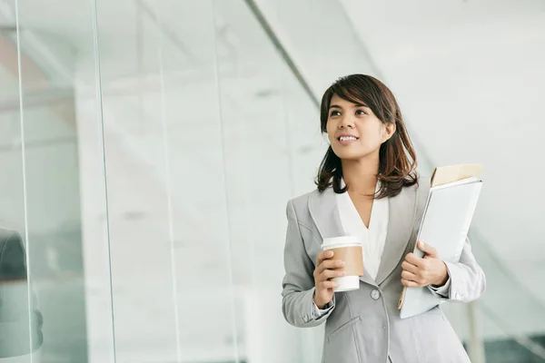 Asian white collar worker with cup of coffee and documents in hands standing in modern office lobby and looking away dreamily.