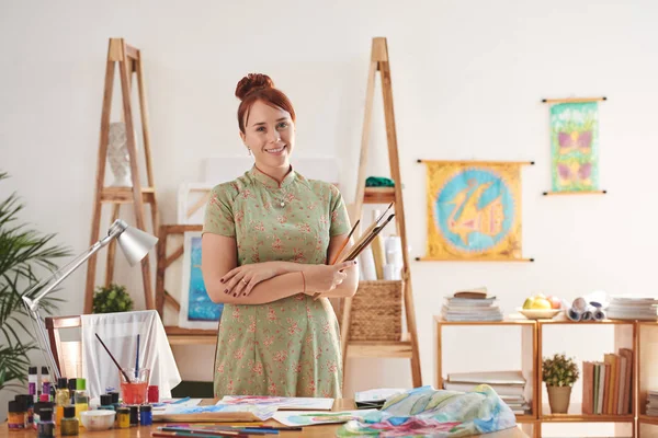 Portrait of happy female art studio owner with paint brushes smiling at camera