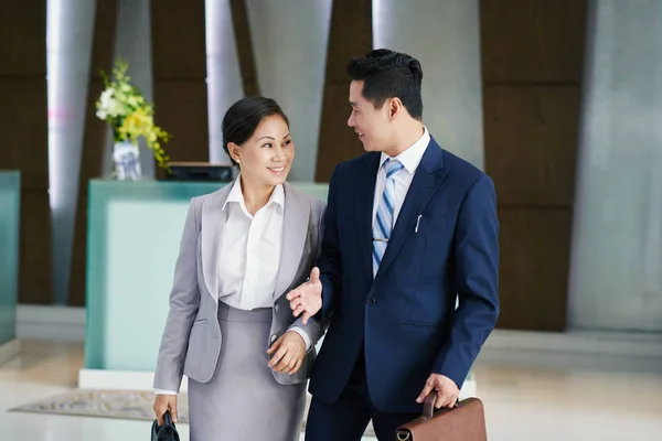 Smiling Asian colleagues in business wear talking while walking in modern office lobby