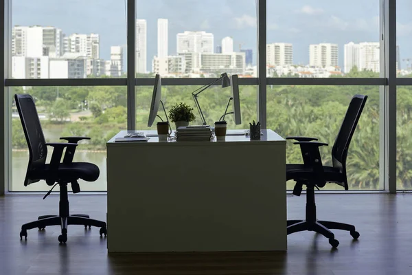 空のオフィスの2つの椅子 コンピュータとコーヒーカップ 背景に大きな都市の景色を望む大きなパノラマの窓があるデスク — ストック写真