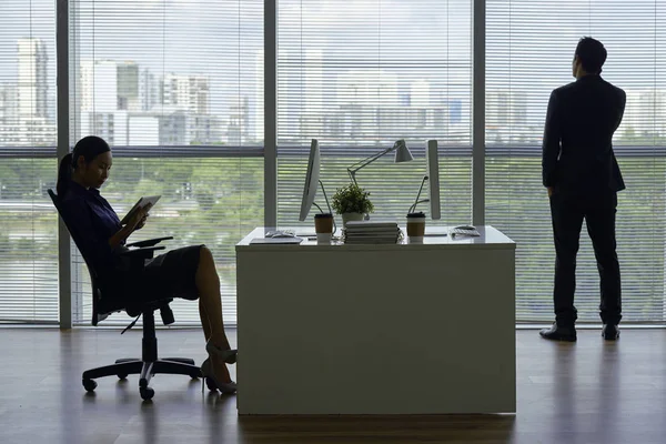 Businesswoman reading information on tablet computer when her coworker talking on phone when looking at big city through panoramic office window