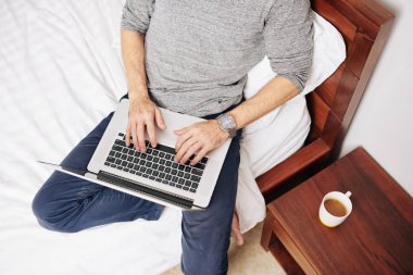 Genç adam bir fincan kahveyle yatakta oturuyor ve laptopta çalışıyor ve programlıyor, yukarıdan bakıyor.