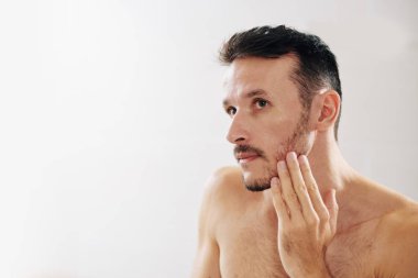 Üstsüz sakallı adam yüzüne dokunuyor ve duş aldıktan sonra aynaya bakıyor.