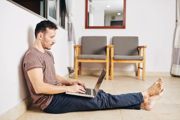 赤脚年轻人坐在敞开的窗户下的地板上 用笔记本电脑编程 — 图库照片