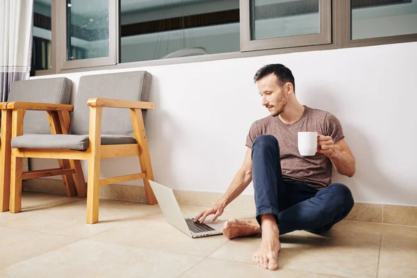 Guapo Joven Descalzo Sentado Suelo Habitación Bebiendo Café Revisando Correos — Foto de Stock